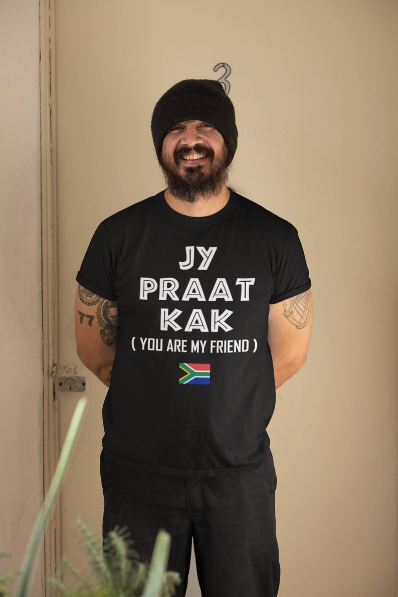 May include: A man wearing a black t-shirt with the text "Jy Praat Kak (You Are My Friend)" and a South African flag. He is smiling and wearing a black beanie.