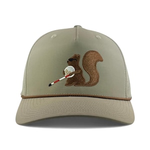 May include: Tan baseball cap featuring an embroidered squirrel wearing sunglasses and holding a golf club. The squirrel is brown with a textured tail. The cap has a brown rope detail and a curved brim.