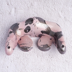 May include: A pink and black climbing hold with a unique, organic shape. The hold features a marbled pattern and two mounting points. The surface has a glossy finish. The hold is designed for rock climbing and bouldering.