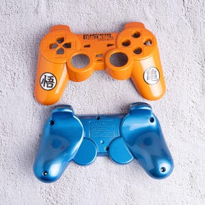 May include: Two game controllers. One is orange with the text "DRAGON BALL" and Japanese characters. The other is blue. Both controllers have buttons and directional pads.