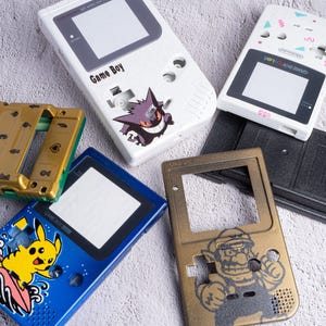 Puede incluir: Una colección de cinco carcasas para consolas portátiles que incluye una carcasa blanca con un gráfico de Gengar, una Game Boy Color blanca, una carcasa dorada con iconos, una carcasa azul con un gráfico de Pikachu y una carcasa dorada con Wario.