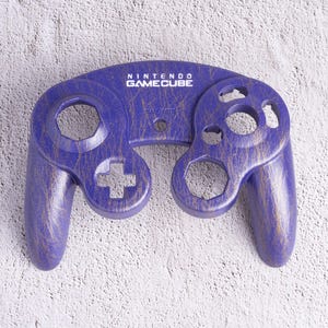 May include: A purple Nintendo GameCube controller with a wood grain finish. The controller has the words "NINTENDO GAMECUBE" printed on the top. The controller has a directional pad, buttons, and analog sticks.