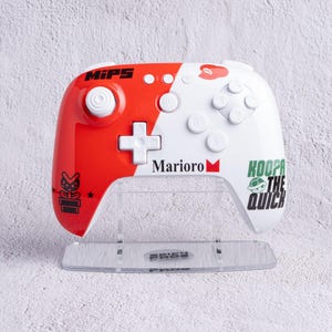 Puede incluir: Un mando de videojuegos rojo y blanco con el texto "MIPS" y "Marioro" impreso. El mando tiene una cruceta direccional y varios botones. Las palabras "Koopa the Quick" están impresas en el lado derecho del mando.