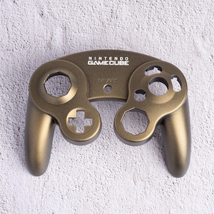 May include: A bronze-colored Nintendo GameCube controller. The controller has the words "NINTENDO GAMECUBE" printed on the top. The controller has a cross-shaped directional pad, a hexagonal button, and several circular buttons.