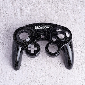 May include: A black Nintendo GameCube controller with a speckled finish. The controller features the GameCube logo and iconic button layout, including a directional pad, analog stick, and action buttons. The controller is designed for gaming.