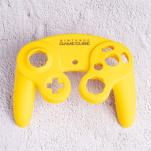 May include: Bright yellow Nintendo GameCube controller. The controller has a distinctive shape with two handles, a directional pad, and several buttons. The text "NINTENDO GAMECUBE" is printed at the top. The controller is made of a smooth material.