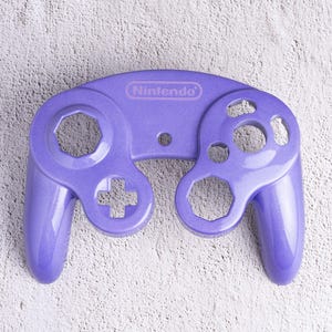 May include: A lavender-colored Nintendo GameCube controller. The controller has a distinctive shape with a central section and two handles. It features a directional pad, analog stick, and various buttons, including the Nintendo logo.