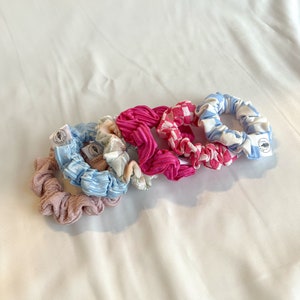 Może przedstawiać: Pięć kolorowych scrunchie z wzorem kwiatowym, różową kratą, różowym, niebieską kratą i jasnoniebieskim. Każde scrunchie ma małą białą metkę z logo.