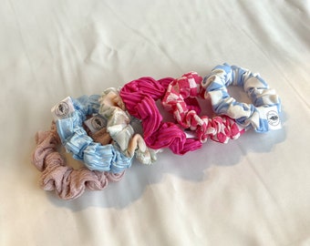 May include: Five colorful scrunchies with a floral pattern, pink gingham, pink, blue gingham, and light blue. Each scrunchie has a small white tag with a logo.