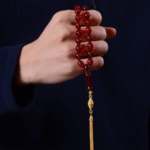 Peut inclure: Un chapelet de prière avec des perles rouge foncé translucides. Les perles sont enfilées avec un séparateur rouge foncé et un gland doré. Les perles sont tenues sur un fond bleu foncé.