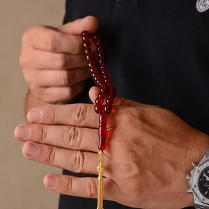 May include: A red prayer bead necklace with a gold tassel. The beads are strung on a cord and the tassel is attached to the end of the cord.