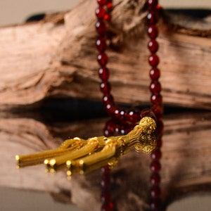 May include: A red beaded prayer necklace with a gold tassel. The necklace is draped over a piece of driftwood.