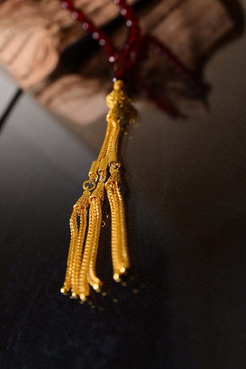 May include: A gold tassel with multiple strands hangs from a red beaded necklace. The tassel is made of intricate gold chain links.