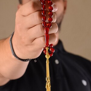 May include: A red prayer bead necklace with a gold tassel. The beads are strung on a cord and the tassel is attached to the end of the cord.