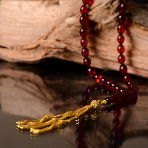 May include: A red prayer bead necklace with a gold tassel. The necklace is resting on a piece of wood.