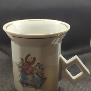 Puede incluir: Taza de porcelana blanca con borde dorado y un diseño de dos campanas azules, una herradura y rosas rosas. La taza tiene un asa cuadrada.