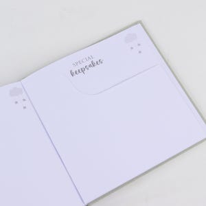 May include: Open baby keepsake book with gray accents. The left page has a cloud and star design. The right page has a pocket with the words "SPECIAL keepsakes" in script. The pages are white.