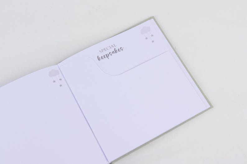 May include: Open baby keepsake book with gray accents. The left page has a cloud and star design. The right page has a pocket with the words "SPECIAL keepsakes" in script. The pages are white.