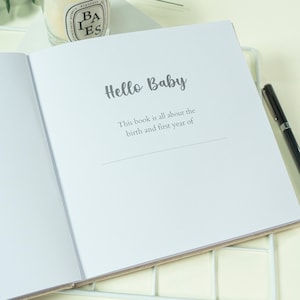 May include: An open baby memory book with the words "Hello Baby" in a script font. The text below reads "This book is all about the birth and first year of". A black pen rests on the right side of the book. The book has a beige cover.