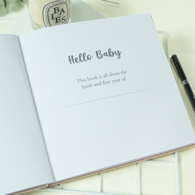 May include: An open baby memory book with the words "Hello Baby" in a script font. The text below reads "This book is all about the birth and first year of". A black pen rests on the right side of the book. The book has a beige cover.