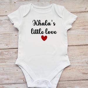 May include: A white short-sleeved baby bodysuit featuring the black text Khala's little love printed on the chest, accompanied by a small red heart icon centered below the lettering. The garment has a round neckline and snap closures.