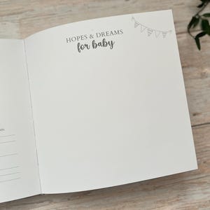 May include: Open baby memory book with the text "HOPES & DREAMS for baby" in gray script. A small banner illustration is in the upper right corner. The book is open to a blank page, ready for journaling.