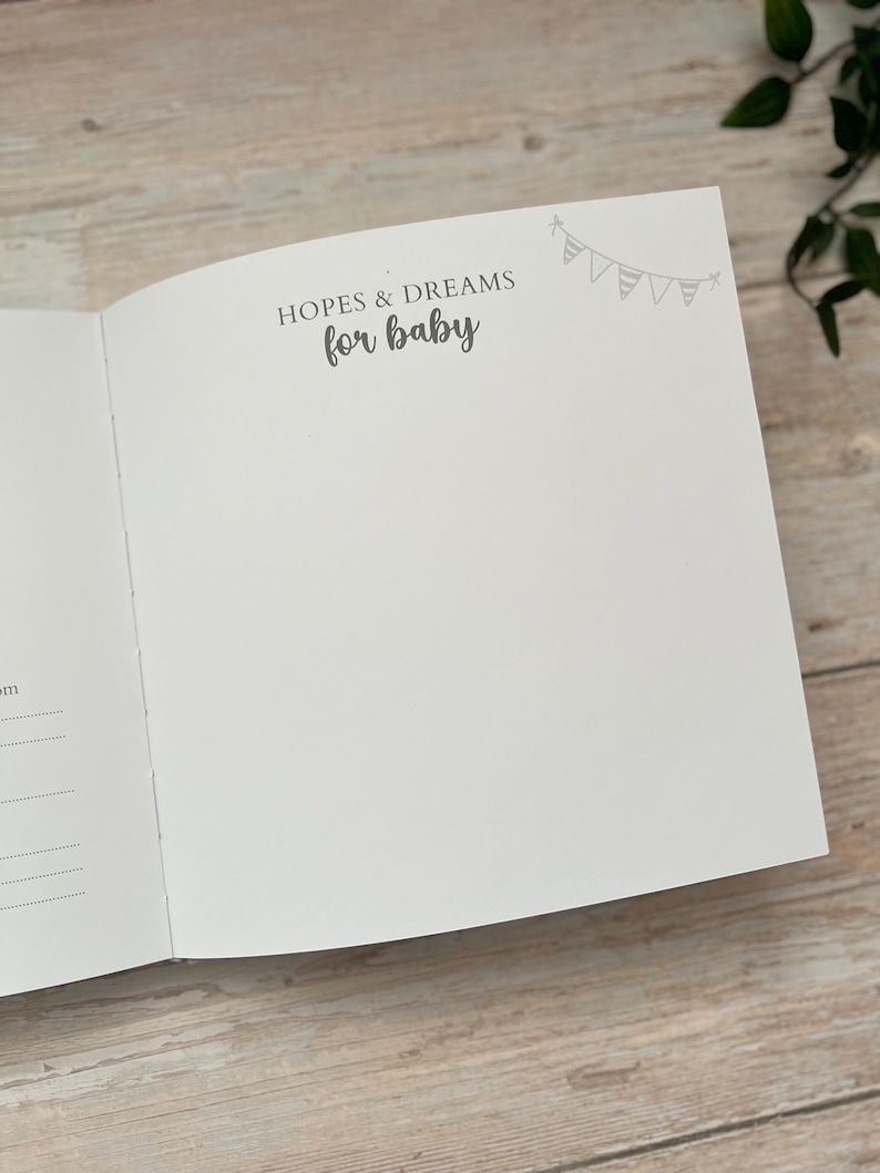 May include: Open baby memory book with the text "HOPES & DREAMS for baby" in gray script. A small banner illustration is in the upper right corner. The book is open to a blank page, ready for journaling.