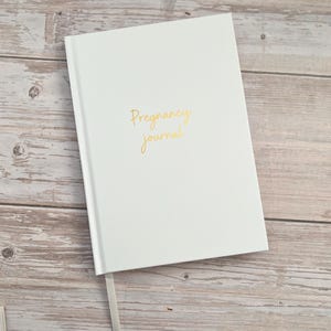 May include: A white hardcover journal with the words Pregnancy journal written in gold cursive script on the front cover. The book features a thin white ribbon bookmark extending from the bottom edge of the spine.