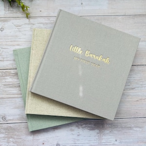 May include: Three hardcover baby memory books in shades of gray, green, and beige are stacked on a wooden surface. The top book is gray with gold lettering that reads "Little Barakah MY FIRST YEAR". The other two books are visible beneath the top book.