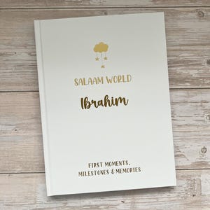 Pode incluir: Um livro branco com um design de nuvem e estrelas douradas. O texto "Salaam World Ibrahim" &eacute; impresso em dourado e marrom. O texto "First Moments, Milestones & Memories" &eacute; impresso em dourado na parte inferior.
