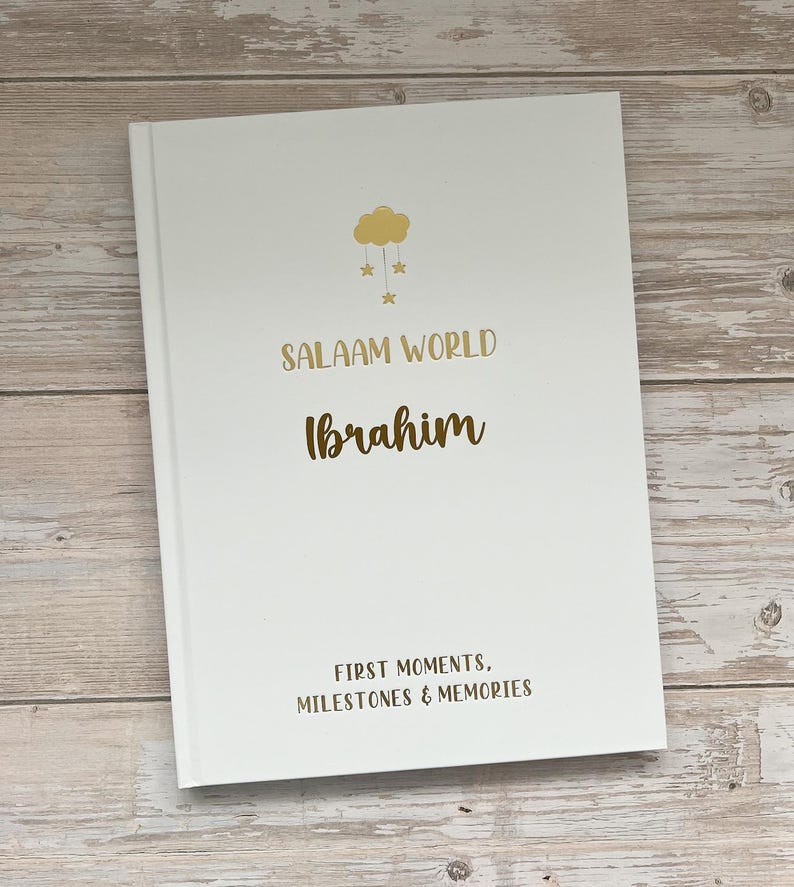 Pode incluir: Um livro branco com um design de nuvem e estrelas douradas. O texto "Salaam World Ibrahim" &eacute; impresso em dourado e marrom. O texto "First Moments, Milestones & Memories" &eacute; impresso em dourado na parte inferior.