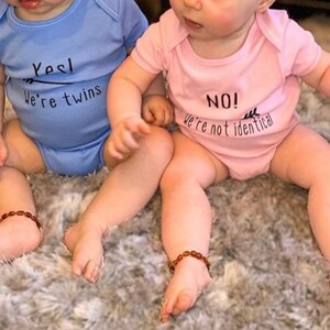 Twins wearing vests, boy twin wearing blue vest with Yes we're twins with arrow pointing to girl twin. Girl twin wearing pink vest with No we're not identical with arrow pointing to boy twin. 100% cotton vests.