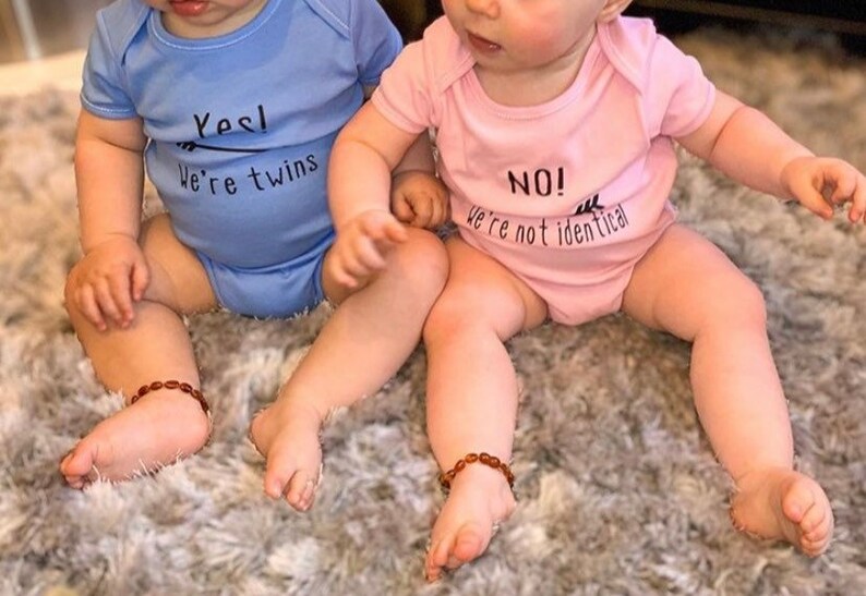 Twins wearing vests, boy twin wearing blue vest with Yes we're twins with arrow pointing to girl twin. Girl twin wearing pink vest with No we're not identical with arrow pointing to boy twin. 100% cotton vests.
