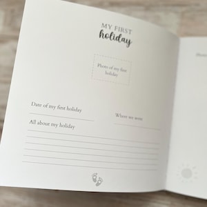 May include: Open baby memory book with the words "MY FIRST holiday" in black script. The book has space for a photo, date, and holiday details. A sun graphic and baby footprints are also visible.