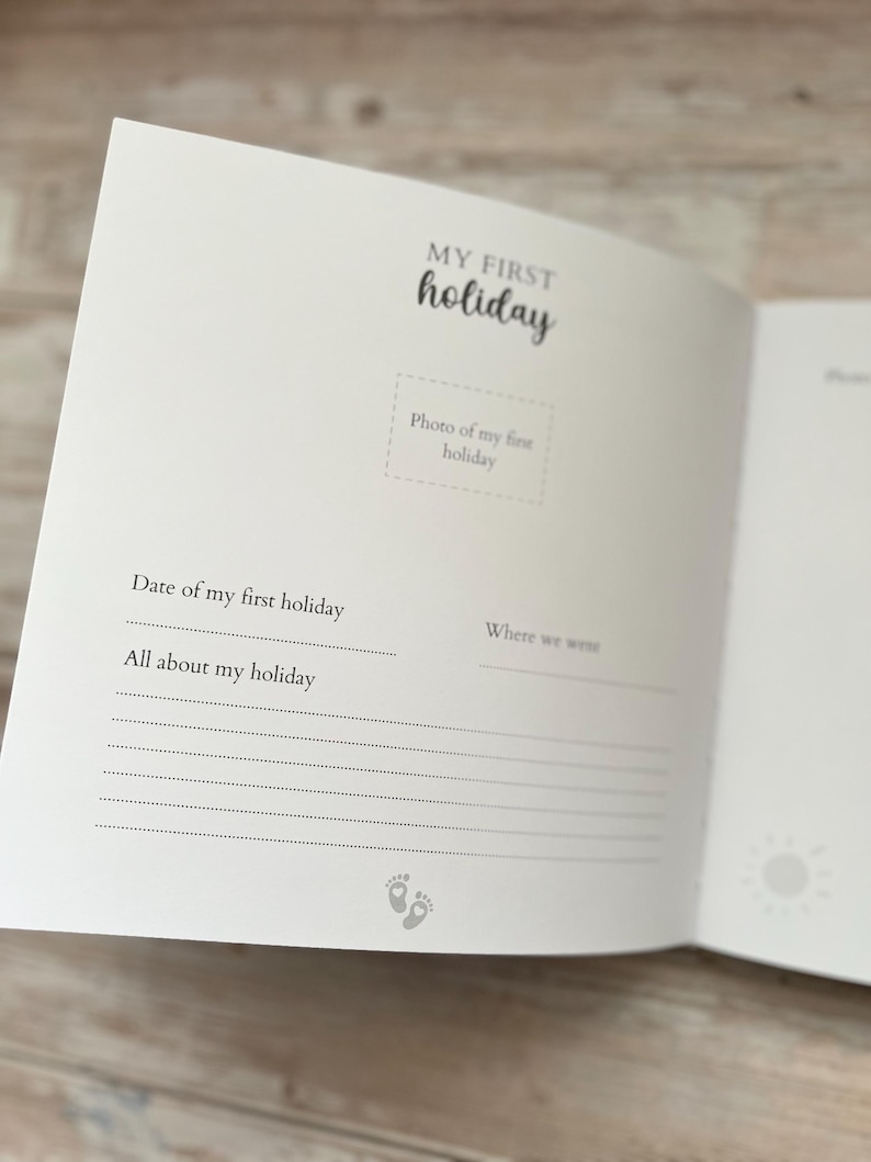May include: Open baby memory book with the words "MY FIRST holiday" in black script. The book has space for a photo, date, and holiday details. A sun graphic and baby footprints are also visible.