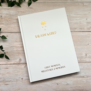 Pode incluir: Di&aacute;rio de capa dura branco com letras douradas que dizem "Salaam World" e "First Moments, Milestones & Memories". A capa apresenta uma ilustra&ccedil;&atilde;o dourada de uma nuvem com gotas de chuva.