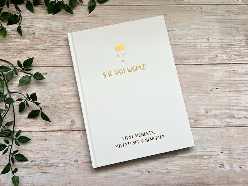 Pode incluir: Di&aacute;rio de capa dura branco com letras douradas que dizem "Salaam World" e "First Moments, Milestones & Memories". A capa apresenta uma ilustra&ccedil;&atilde;o dourada de uma nuvem com gotas de chuva.