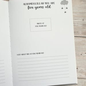 Pode incluir: Um livro branco com um design de nuvem e estrelas em preto e cinza. O texto "ALHAMDULILLAH YOU ARE five years old" est&aacute; impresso na capa. O livro tem um espa&ccedil;o para fotos com o texto "PHOTO AT FIVE YEARS OLD" e uma se&ccedil;&atilde;o para escrever "A BIT ABOUT YOU AT FIVE YEARS OLD".