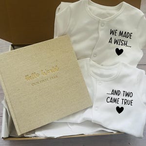 May include: A gift box containing a beige fabric-covered memory book with the gold embossed text Hello World OUR FIRST YEAR and two white long-sleeved baby bodysuits with black text reading WE MADE A WISH... and ...AND TWO CAME TRUE.