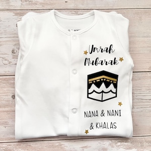 May include: White baby onesie with gold text that reads "Umrah Mubarak" and a black and gold illustration of the Kaaba. The onesie also has gold stars and the text "Nana & Nani & Khalas".