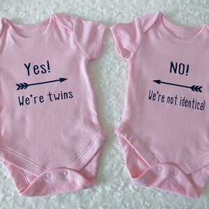 Two pink baby vest or bodysuits, one with Yes we're twins with arrow pointing to the other vest. Other with No we're not identical with arrow pointing to other vest. Arrows are in between the text. All black design. Customisation available