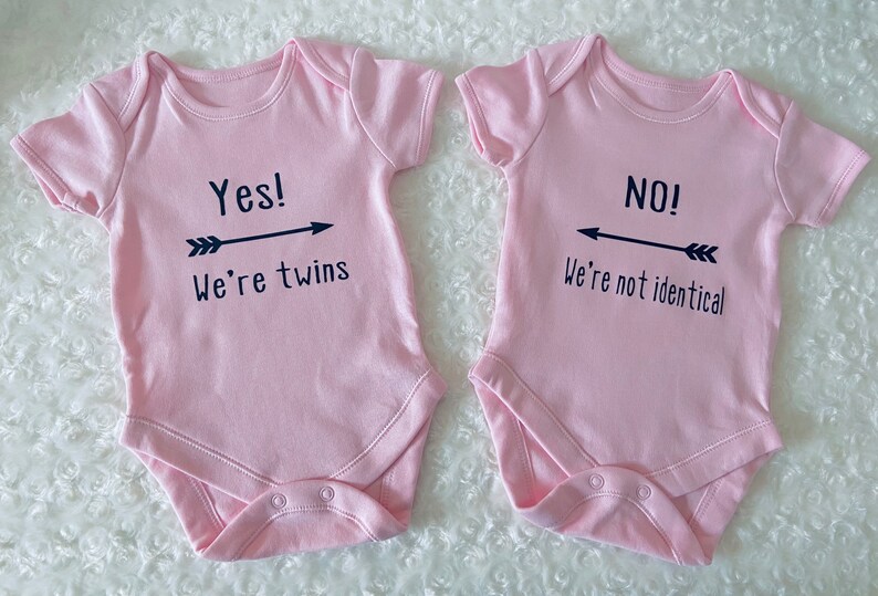Two pink baby vest or bodysuits, one with Yes we're twins with arrow pointing to the other vest. Other with No we're not identical with arrow pointing to other vest. Arrows are in between the text. All black design. Customisation available