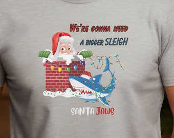 May include: A light gray t-shirt featuring a Christmas-themed graphic. The design includes Santa Claus in a chimney, a shark with Christmas lights, and the text "WE'RE GONNA NEED A BIGGER SLEIGH" and "SANTA JAWS".