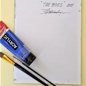 May include: A white canvas with a blue border. The canvas is signed "THE BOSS" 2021, with the artist's signature below. There is a blue tube of acrylic paint and two paintbrushes on the canvas.
