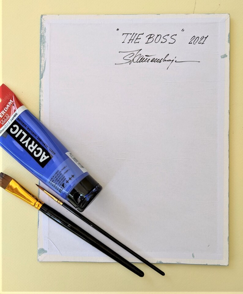 May include: A white canvas with a blue border. The canvas is signed "THE BOSS" 2021, with the artist's signature below. There is a blue tube of acrylic paint and two paintbrushes on the canvas.