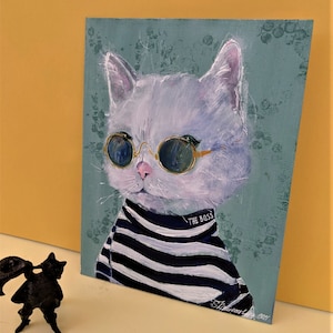 May include: A painting of a white cat wearing round sunglasses and a black and white striped sweater. The cat is looking directly at the viewer. The text "THE BOSS" is written on the sweater. The background is a light blue-green color with a floral pattern.