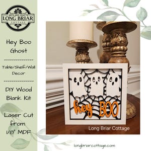 May include: A square wooden sign featuring three white ghost cutouts layered over a black spiderweb design. The orange text reads "hey BOO" at the bottom. The sign is framed in white and is laser cut from 1/8 inch MDF material for DIY assembly.