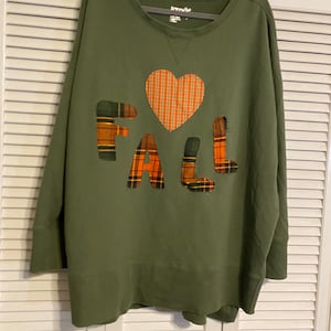 Op de afbeelding: Een olijfgroen sweatshirt met lange mouwen en een geruite hartapplicatie boven het woord FALL, waarbij elke letter ook is gemaakt van oranje, groene en zwarte geruite stoffen patches met zichtbare gerafelde randen.
