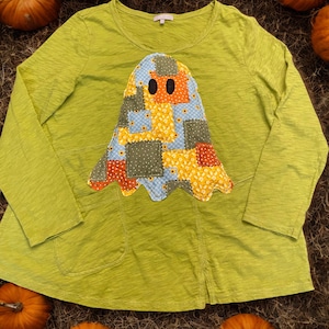 May include: A long-sleeved, lime green shirt with a patchwork ghost applique. The ghost is made of various fabric patterns in blue, yellow, orange, and green. The shirt has a round neckline and two front pockets. Pumpkins surround the shirt.