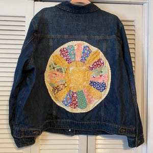 Op de afbeelding: Een donkerblauwe spijkerjas met een groot, kleurrijk, cirkelvormig patchwork-ontwerp op de rug. Het patchwork bevat verschillende bloemen- en patronenstoffen in tinten geel, roze, blauw en groen. De jas heeft een klassieke kraag en knoopsluitingen.